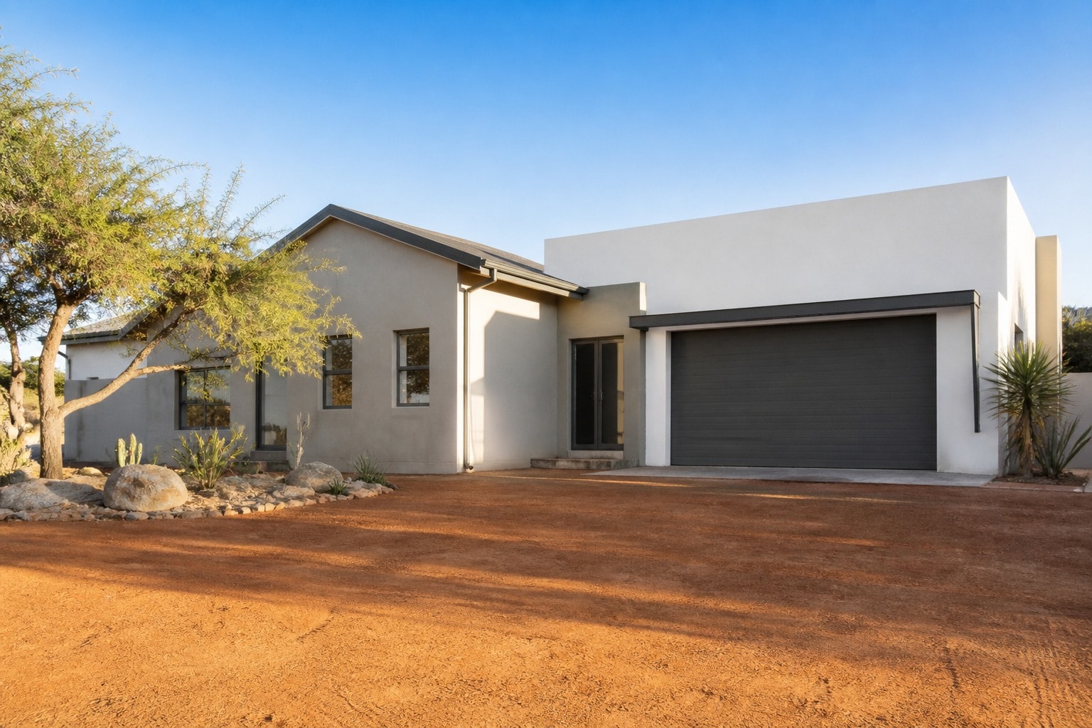 Single-storey house with a grey plastered living wing, white garage block, dark roller door, acacia tree, and a wide red-earth driveway under a clear blue sky